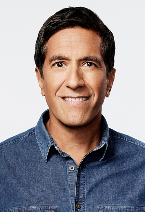A man with neatly trimmed dark hair poses in a dark chambray shirt in front of a stark white background.