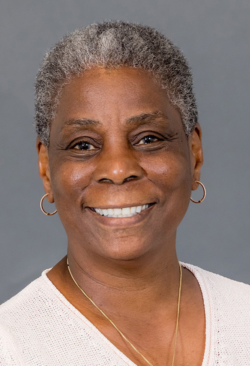 Woman with graying short hair smiles broadly in front of a gray studio backdrop. She's wearing a gray jacket and black blouse and wears silver hoop earings and necklace.