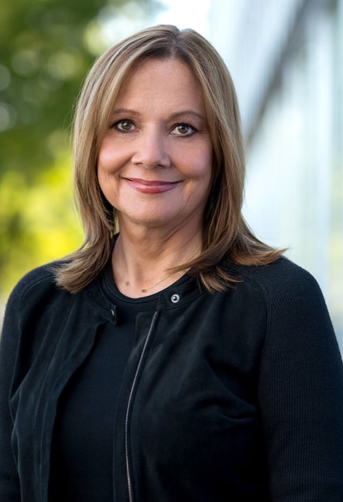  A woman with shoulder length hair smiles broadly and is wearing a smart black jacket and is outside with soft focus trees and a building in the background