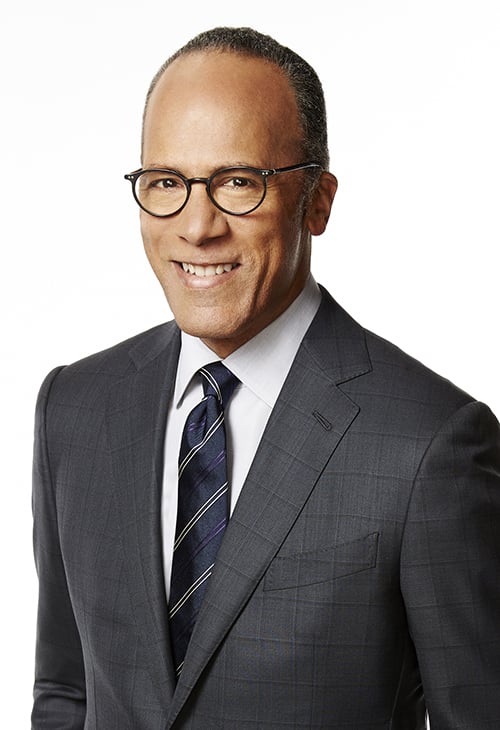 A man dressed in a gray suit jacket wears a navy tie with white stripes and rounded eyeglasses.